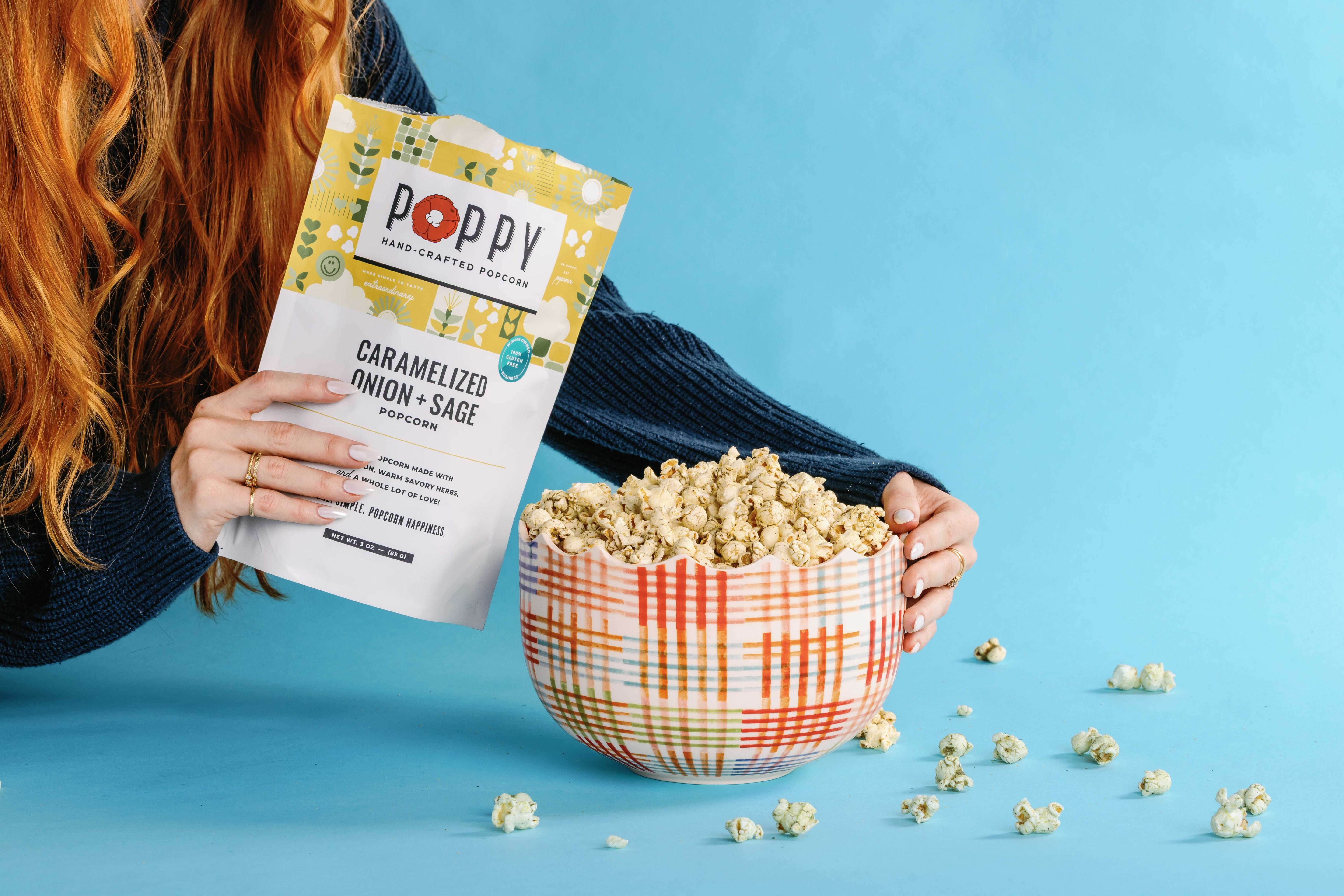 A person with red hair holds a bag of Poppy Handcrafted Popcorn next to a plaid bowl filled with popcorn on a bright blue background.